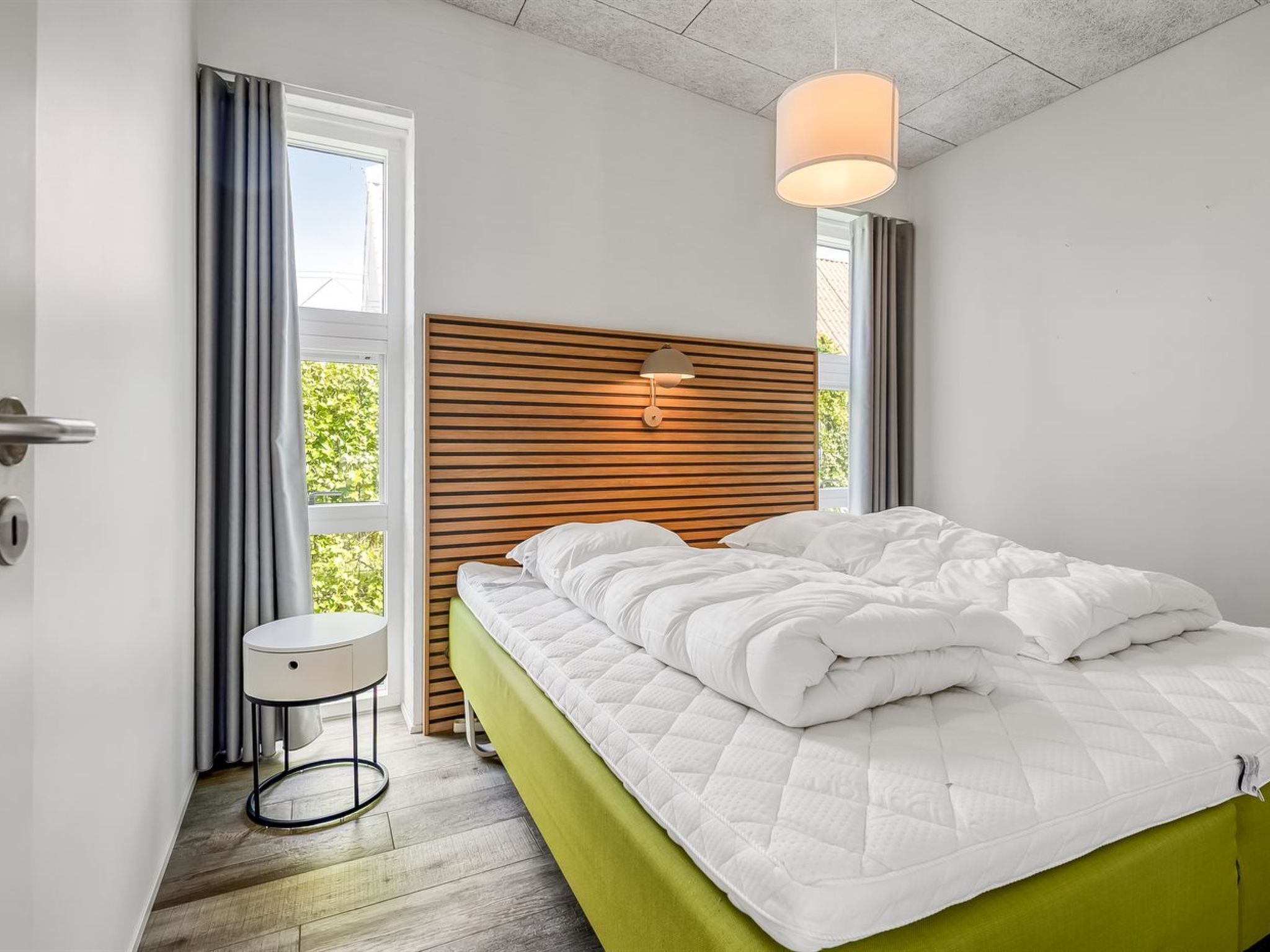 A modern bedroom with a wooden headboard, a bedside table, a green bedspread, and a hanging light fixture.