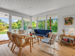 A spacious room with a large window, a blue sofa, wicker chairs, a rocking chair, and a small table with a lamp and decorative items.