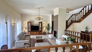 A spacious living room with a large TV, comfortable seating, a staircase, and a chandelier.