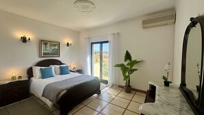 A bedroom with a bed, a nightstand, a potted plant, a mirror, and a view of the outside.