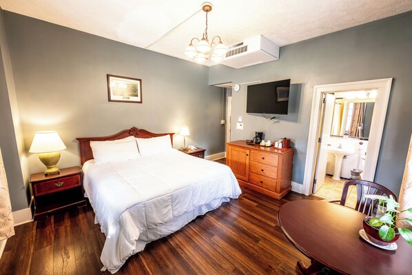 A hotel room with a large bed, a wooden dresser, a desk, a chair, a television, and a bathroom visible through an open door.