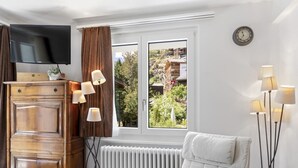 A room with a TV, a wooden cabinet, a radiator, a white chair, and a window with curtains. - Zermatt, 4 pers/3 lits - Balcon avec vue & jardin (Zermatt)