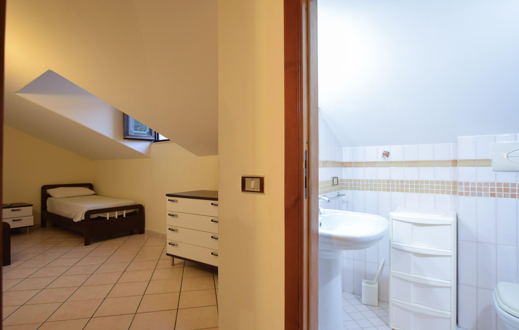Ein Schlafzimmer mit einem Bett, einer Kommode und einem kleinen Schreibtisch. Durch eine offene Tür ist ein Badezimmer mit Waschbecken und Toilette zu sehen.