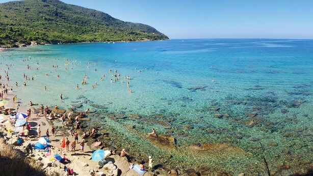 Ein Strand mit klarem türkisblauem Wasser, Felsen und einer Hügelkette im Hintergrund.