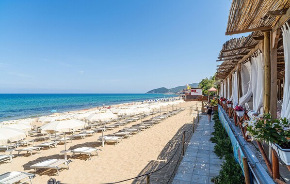 A beach with rows of sunbeds and umbrellas, a clear blue sky, and a building with a bamboo roof.