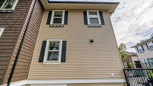 Een woonhuis met twee verdiepingen, bekleed met beige panelen en zwarte luiken, met een klein balkon en een zichtbare schoorsteen.