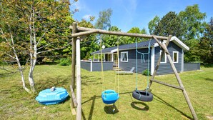 A wooden swing set with two swings and a tire swing, a blue plastic tub, and a house in the background.