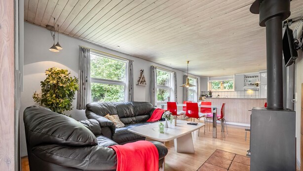 A modern living room with a black leather sofa, a white coffee table, and a red throw blanket. There is a dining area with red chairs and a kitchen with a black stove. - 3 bedroom stunning home in Tarm (Tarm)