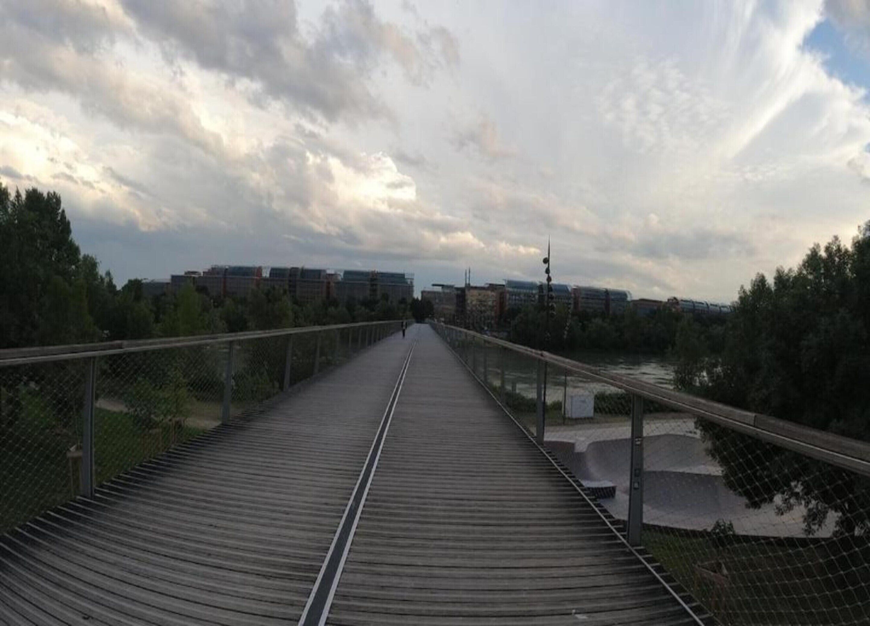 Un long pont piétonnier rectiligne, doté d’une rambarde métallique et d’une passerelle en bois, qui mène vers une étendue d’eau et des bâtiments situés au loin.