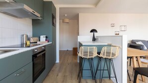 A modern kitchen with a built-in oven, a stove, and a stainless steel range hood. There is a bar area with two stools and a small island with a bar stool. The kitchen has a light-colored backsplash and dark green cabinets. - Dify Republique - Villeurbanne (Villeurbanne)