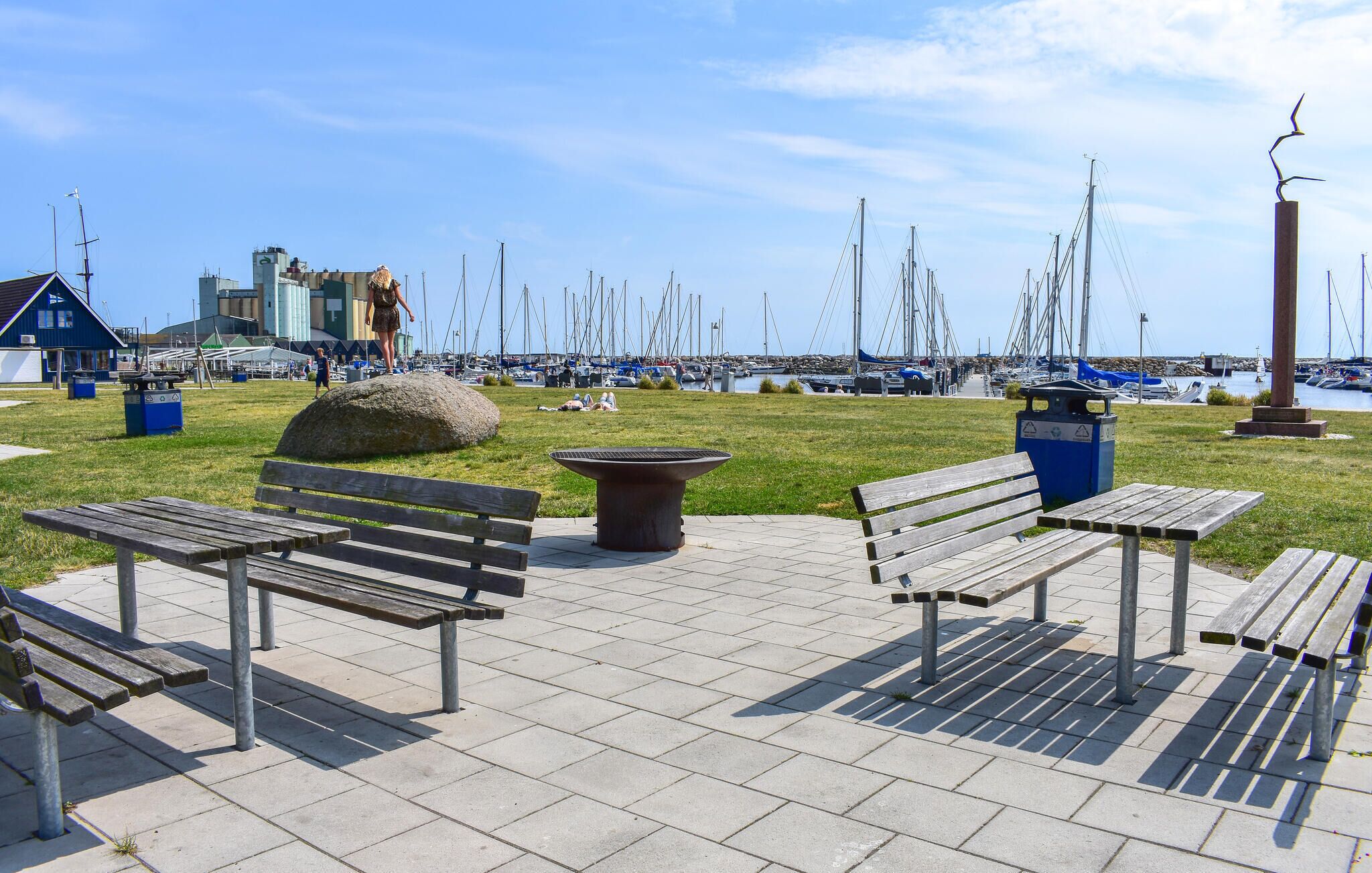 A waterfront park with benches, a large rock, and a marina in the background.