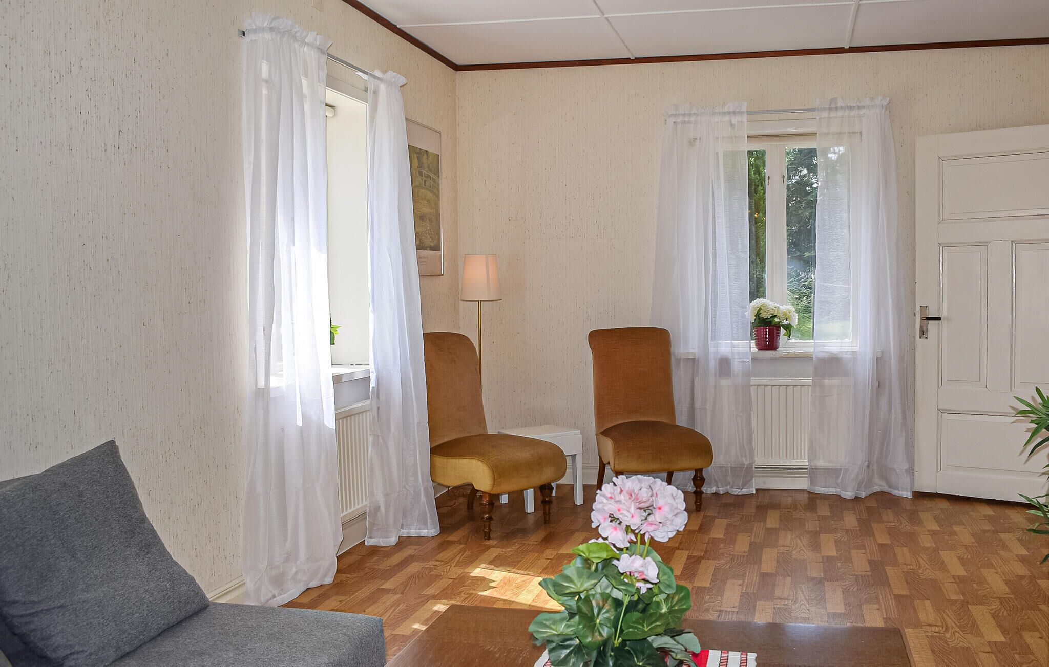 A room with two brown armchairs, a grey sofa, a wooden floor, and a window with white curtains.