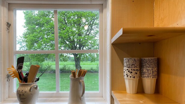 A kitchen window with a view of a green landscape, a white mug with utensils, and two patterned cups on a wooden shelf. - Cozy home in Kalmar with WiFi (kalmar)