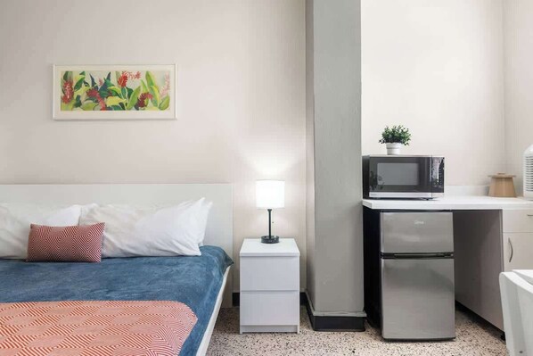 A hotel room with a bed, a nightstand with a lamp, a microwave, a refrigerator, and a small potted plant.