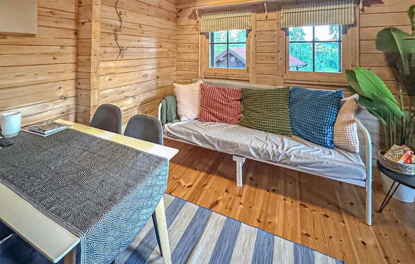 A cozy cabin interior with a wooden interior, a sofa with colorful cushions, a dining table with chairs, and a window with a view of trees.