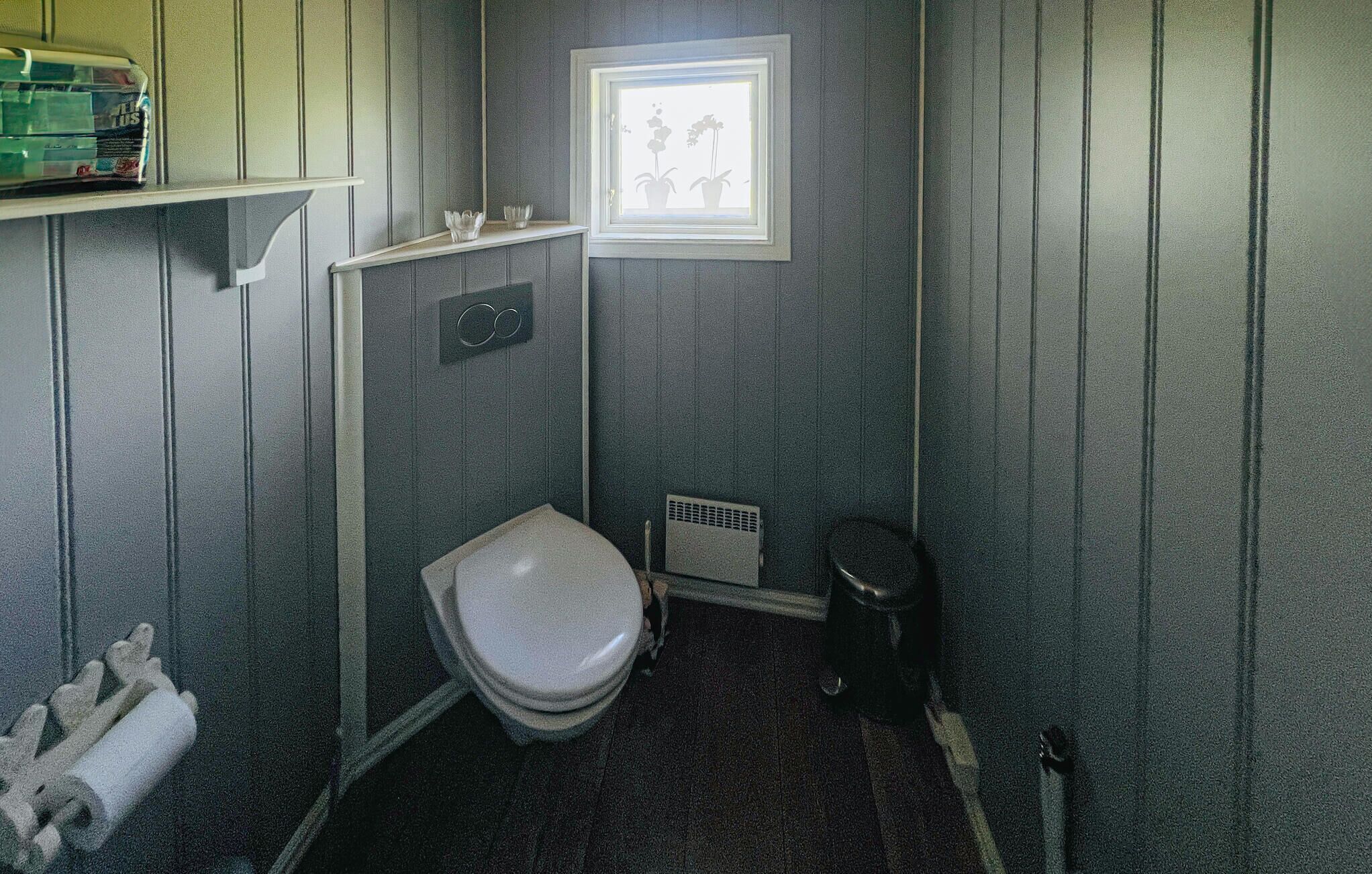 A compact bathroom with a toilet, a small shelf with toiletries, a vent, and a framed picture on the wall.