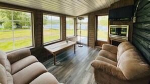 Une pièce chaleureuse avec un plancher en bois, de grandes fenêtres donnant sur un lac, une télévision et un banc.