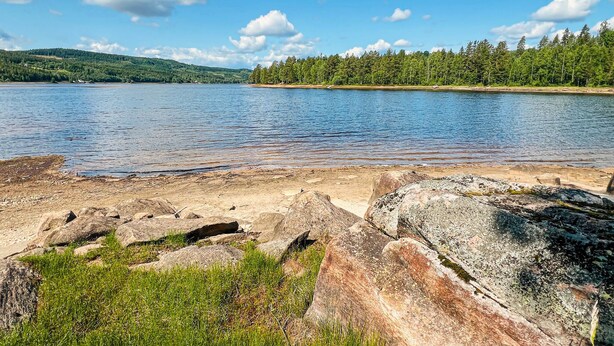 Une vue sur un lac, avec des rives rocheuses, une eau claire et une colline boisée en arrière-plan.
