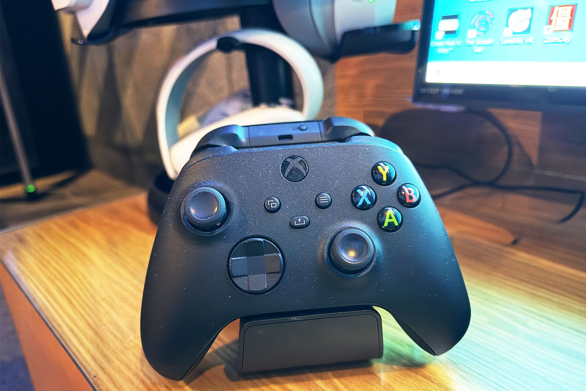 A black gaming controller on a wooden surface with a monitor displaying game icons in the background.