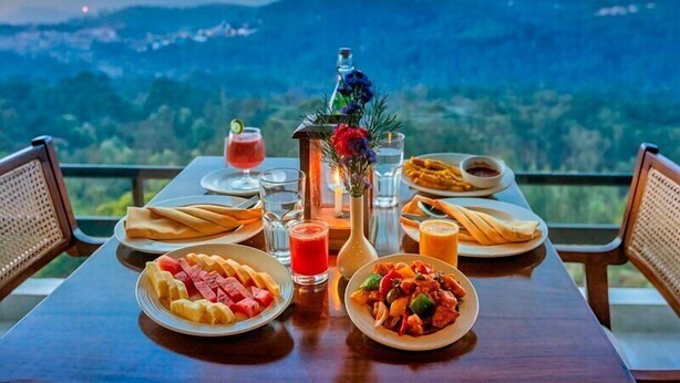 A dining table set with plates of food, drinks, and a decorative centerpiece, overlooking a mountainous landscape. - Sleeping Beauty By Nature Resorts  (Aruvankadu)