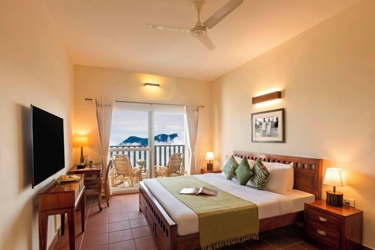A hotel room with a bed, a desk, a chair, a TV, and a balcony with a view of the mountains.