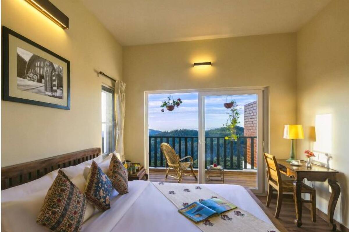 A hotel room with a large bed, a balcony with chairs, and a table with a lamp.