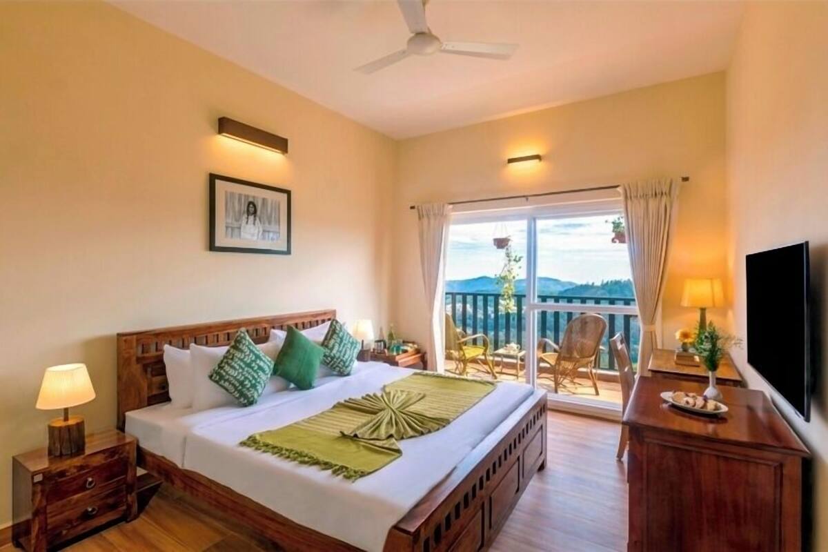 A hotel room with a bed, a TV, a balcony with chairs, and a view of the landscape.