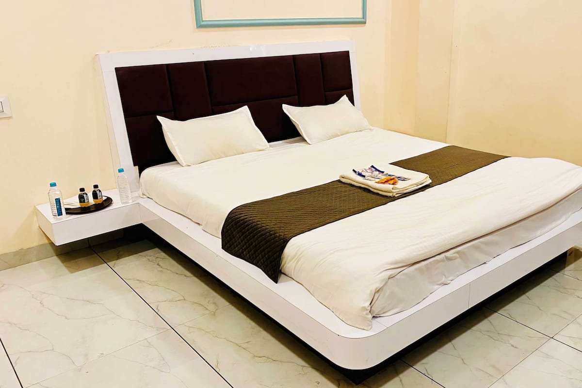 Een netjes opgemaakt bed met een wit-bruine sprei, een kussen en een gevouwen tijdschrift. Er staat een klein nachtkastje met een fles en een zak tissues.