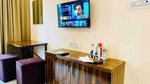 A hotel room with a wooden desk, a television mounted on the wall, a power outlet, and a small table with a remote control and bottles. - Hotel Kush Palace (Ujjain)