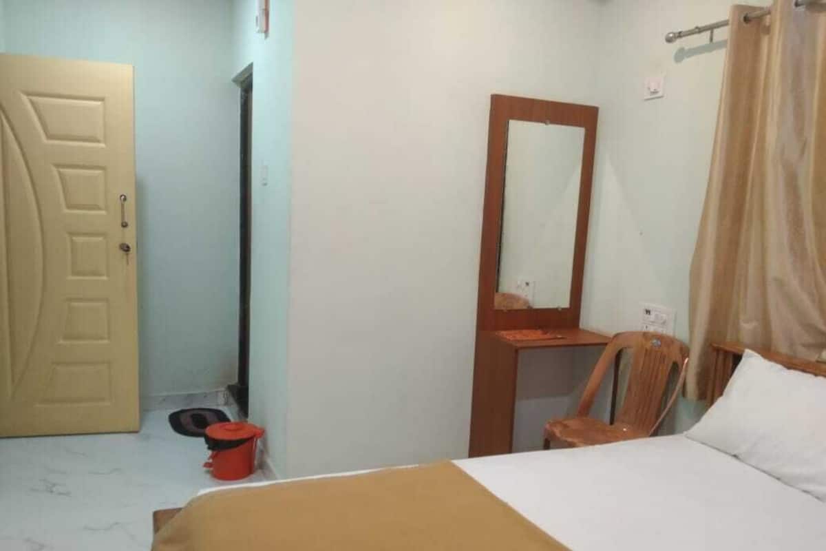 Un dormitorio con una cama, una silla, un escritorio de madera, un espejo, una puerta y una cortina.