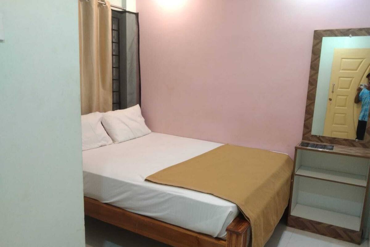 Una cama individual con estructura de madera, una colcha color beige y ropa de cama blanca. Un espejo cuelga en la pared a la derecha, reflejando una puerta.
