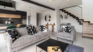 Een moderne woonkamer met een grijze bank, een bijzettafeltje en een trap met een zwarte leuning.