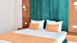 Una cama bien tendida con ropa de cama blanca y naranja, un cabecero tapizado en color verde azulado y mesitas de noche de madera con lámparas fijadas a la pared.
