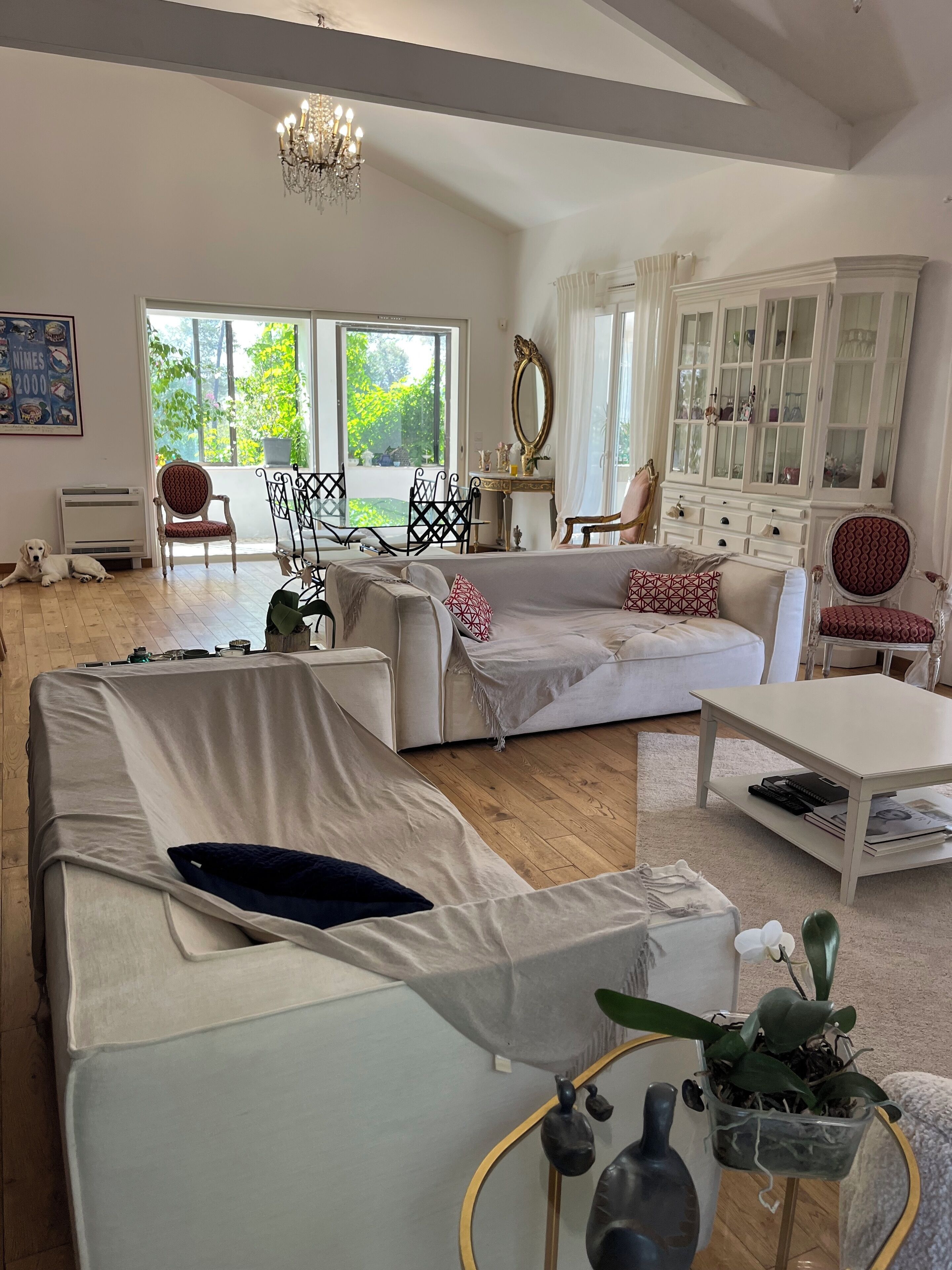 A spacious living room with a large sofa, a coffee table, and a dining area with chairs. There is a chandelier, a cabinet with glass doors, and a window with a view of greenery.