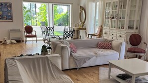 A spacious living room with a large sofa, a coffee table, and a dining area with chairs. There is a chandelier, a cabinet with glass doors, and a window with a view of greenery.