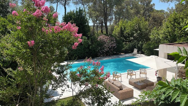 A pool area with a clear blue pool, surrounded by lush greenery and trees. There are wicker lounge chairs and a white outdoor table.