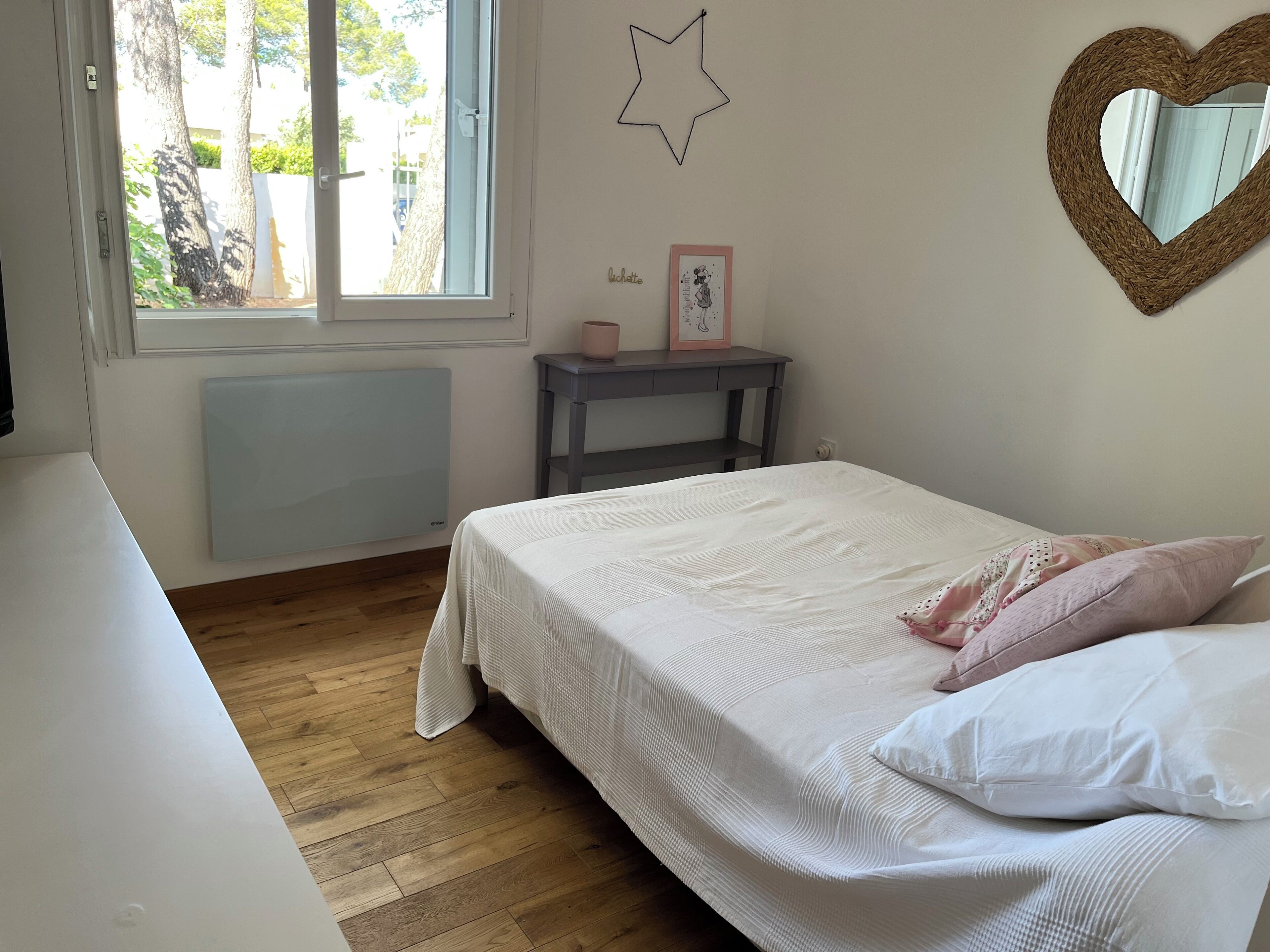 Un dormitorio con cama, mesita de noche, adorno en la pared con forma de estrella, espejo con forma de corazón y ventana con vista a árboles.