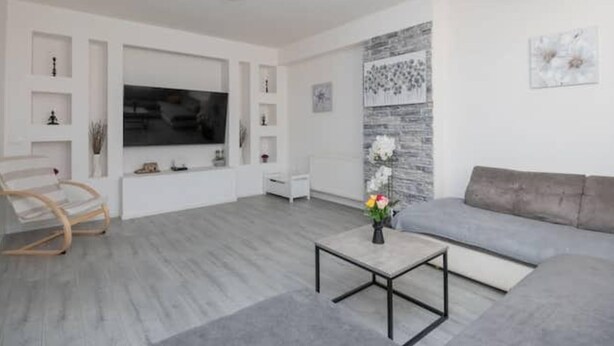 Ein modernes Wohnzimmer mit grauem Sofa, einem Holzstuhl, einem Couchtisch mit einer Vase und einem großen Flachbildfernseher an der Wand.