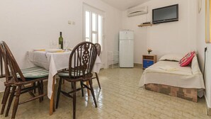 A small, well-lit room with a dining table set for two, a bed with a red pillow, a white refrigerator, and a mounted TV. - Charming Apartments Dokoza Vir (Vir)