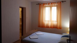 A small, single-bed room with a window covered by orange curtains, a door to another room, and a bedside table with a pillow and a book. - Charming Apartments Dokoza Drage (Pakostane)