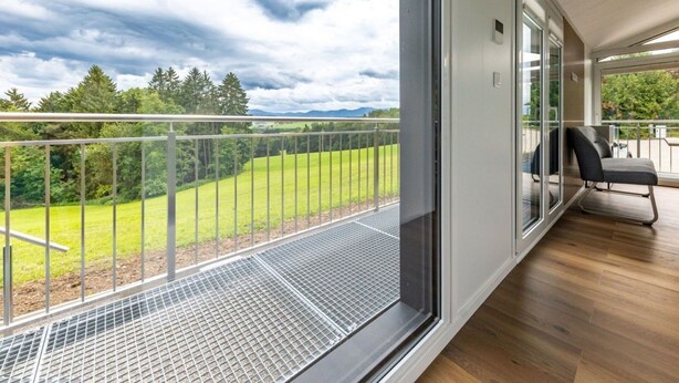 Een balkon met een metalen hekwerk, een stoel, en uitzicht op een groen veld en bomen onder een bewolkte lucht.