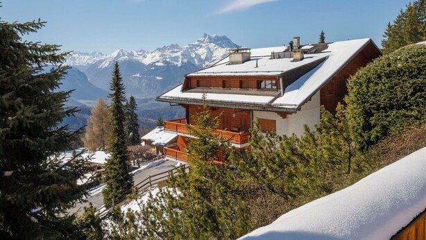 Un chalet de montagne doté d’un toit en pente, d’une façade en bois et offrant une vue sur des montagnes enneigées.