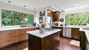 A modern kitchen with wooden cabinets, a central island, and a large window overlooking greenery. - Family Retreat -Mt Rainier Views • Pool • Hot Tub • Sleeps 11 (Auburn)