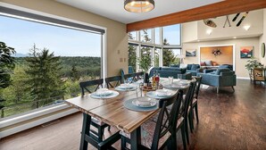 A spacious dining area with a wooden table set for four, large windows offering a view of the forest, and a cozy living area with blue sofas and a wooden ceiling beam. - Family Retreat -Mt Rainier Views • Pool • Hot Tub • Sleeps 11 (Auburn)
