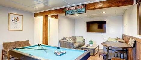 A pool table with a billiard cue, a sofa, a bar area with stools, and a television mounted on the wall.