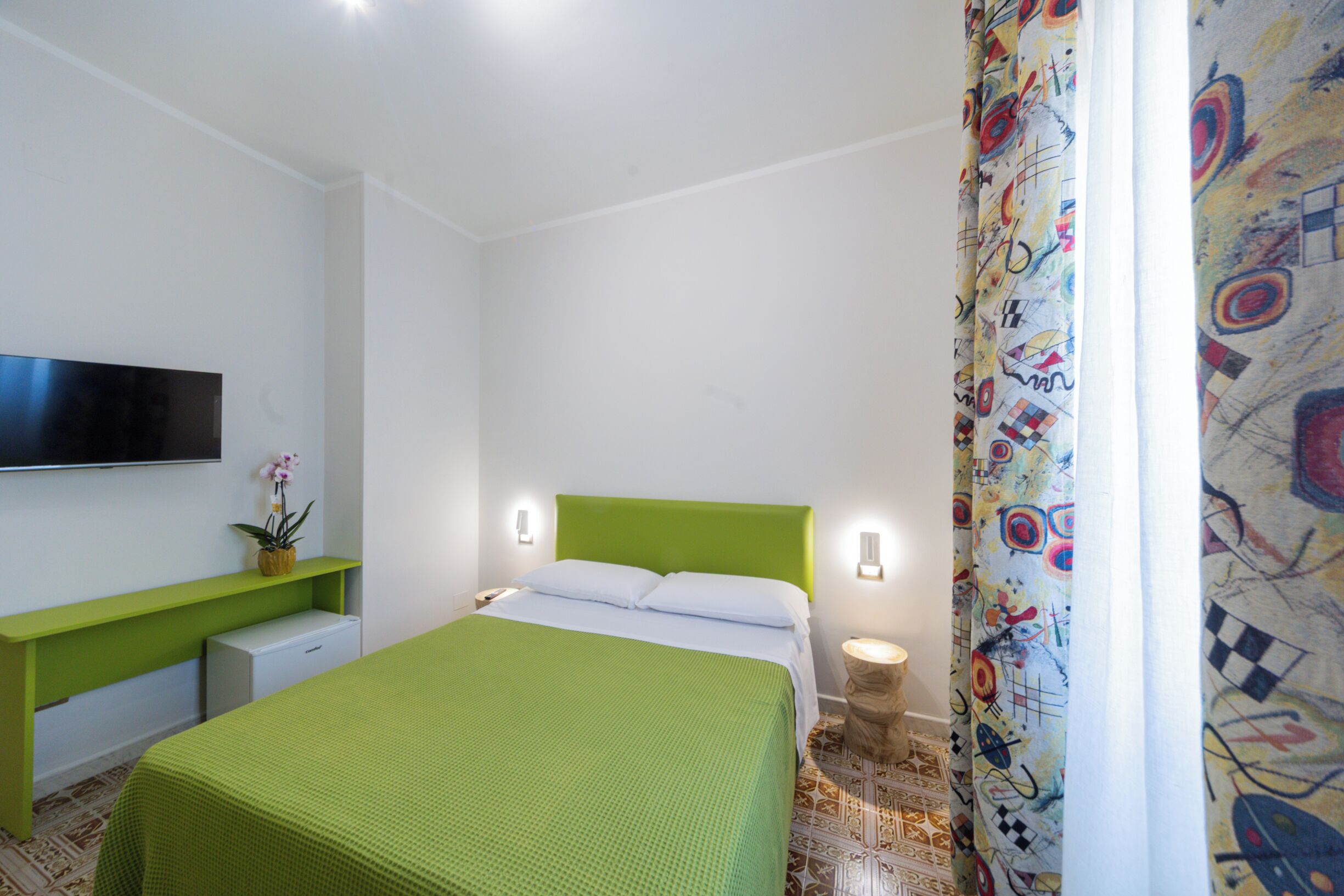 A bedroom with a green bed, a flat-screen TV on the wall, a bedside table, and a small bedside lamp.