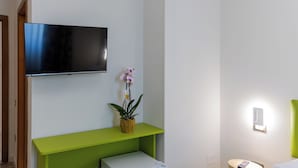 Habitación de hotel con televisión de pantalla plana montada en la pared, una consola de mesa verde, una orquídea en maceta y una mesita de noche con lámpara.