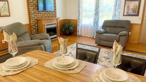 A dining area with a wooden table set for four, two grey armchairs, a brick fireplace, and a large window with curtains. - Lydia's Lodge in Pelham (Pelham)