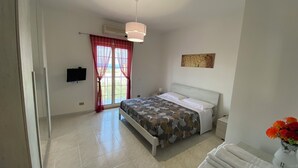 Een slaapkamer met een bed, nachtkastje, televisie, raam met gordijnen, plafondlamp en een aan de muur hangend kunstwerk.