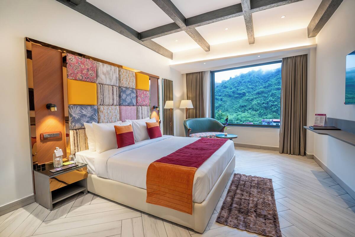 Een moderne hotelkamer met een groot bed, een nachtkastje, een bureau, een stoel en uitzicht op een bebost gebergte.